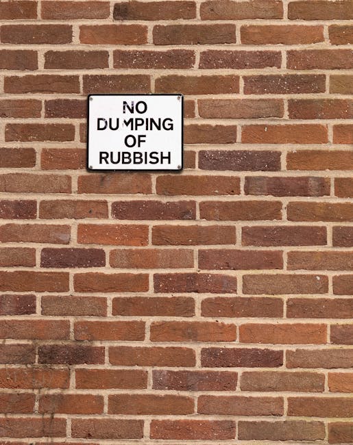 A rectangular white sign with black lettering mounted on a red brick wall. The sign reads 'NO DUMPING OF RUBBISH' with the words 'NO' and 'OF' slightly misaligned, and some letters partially obscured or cut off, suggesting wear or improper installation. The brick wall features a standard running bond pattern with reddish-brown and darker brown bricks, mortar joints visible between them. The surface of the bricks is textured with some variation in colour, and the lighting appears natural, casting soft shadows beneath the sign and on the wall surface. The image captures a section of the exterior wall, possibly part of a building facade, serving as a visual reminder against illegal rubbish disposal, aligning with the theme of waste management and private waste collection services provided by Waste Disposal Twickenham.