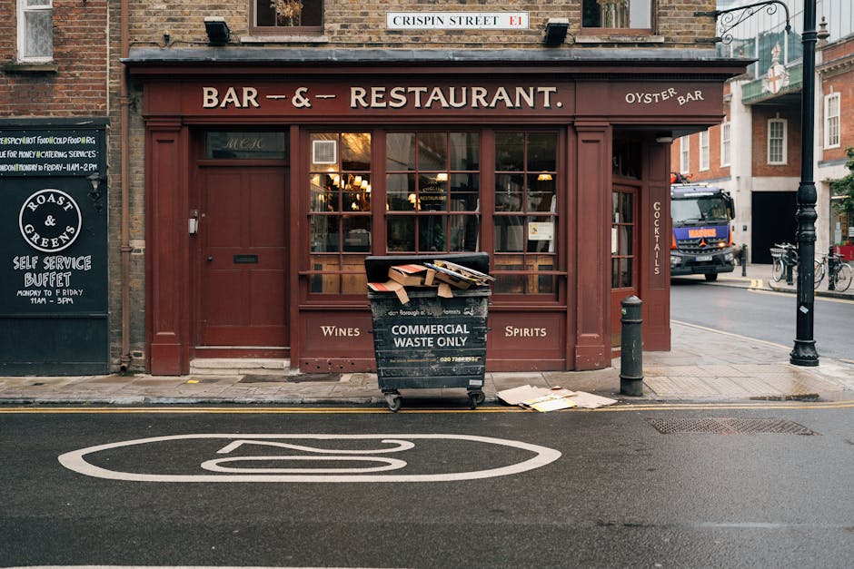 A black wheeled bin labeled 'COMMERCIAL WASTE ONLY' in white text, positioned in front of a café or restaurant with a red-brown exterior. The bin is filled with flattened cardboard boxes and paper waste, some leaning out over the top. The storefront features large glass windows with warm lighting inside, and signage indicating wines, spirits, and cocktails. To the left, a black chalkboard sign advertises a self-service buffet, while on the right, a black bollard and a lamppost are situated on the sidewalk. The scene is set on a paved street with a marked parking space in the foreground and a street with vehicles, including a blue truck, visible in the background. The overall environment suggests an urban area where private waste storage and collection may be part of routine rubbish removal practices managed by Waste Disposal Twickenham, supported by private or independent waste handling services.