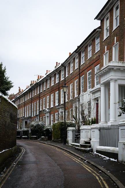 A row of five-story red brick residential buildings with white decorative trim and columned entrances, situated along a narrow, curved street in an urban area. The buildings feature numerous white-framed sash windows, some with small balconies or window ledges, and the façades are lined with chimneys on the roof. In the foreground, there is a pavement with a low brick wall on the left and a white-painted boundary wall with a small set of steps leading to one of the property entrances. The street is damp, possibly from recent rain or snowmelt, and the sky appears overcast, creating a muted, grey lighting atmosphere. A black street lamp is positioned between two doorways, and sparse, leafless shrubs or small trees grow along the front of the buildings, some with remnants of winter snow on the ground or plant branches. This scene reflects a residential street suitable for private waste collection or alternative rubbish services, which Waste Disposal Twickenham could support for local properties.