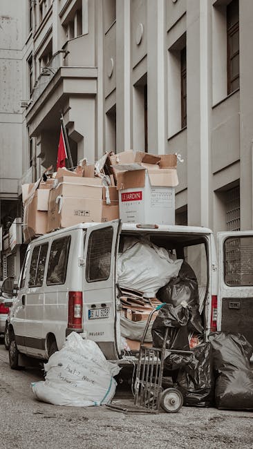 A white panel van with its rear doors open, parked on a city street in front of a beige multi-storey building with vertical architectural features and small windows. The van is filled with various types of waste including large black garbage bags, cardboard boxes, and wooden pallets, some of which are stacked inside and some spilling onto the pavement. Additional waste bags and discarded materials are placed on the ground next to the vehicle, including a small hand trolley without contents and a few loose cardboard pieces. Cardboard boxes of different sizes and textures, some flattened, are visible both inside and on top of the van, with one marked with the brand 'Leardom'. The scene suggests a private waste collection or on-site clearance service, such as those provided by Waste Disposal Twickenham, handling rubbish removal in an urban environment. The lighting appears natural, capturing the details of the waste and surrounding architecture clearly, emphasizing the context of rubbish clearance operations in a city setting suitable for professional waste management services.