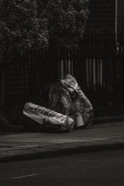 Two large black plastic trash bags filled with waste are positioned on a sidewalk near the curb in front of a black metal fence with vertical bars and lattice panels. The bags appear crumpled and bulky, with some visible text and markings on their surfaces. Behind the fence, dense leafy greenery extends upwards, partially obscuring a structure or building in the background, which is not fully visible. The scene is outdoors, with natural lighting creating slight shadows and highlighting the textures of the plastic bags and the street surface. The bags are likely placed temporarily for rubbish collection or private waste disposal, consistent with the context of rubbish removal services offered by Waste Disposal Twickenham. The environment appears residential or semi-urban, with an emphasis on the collection point for waste outside a property.